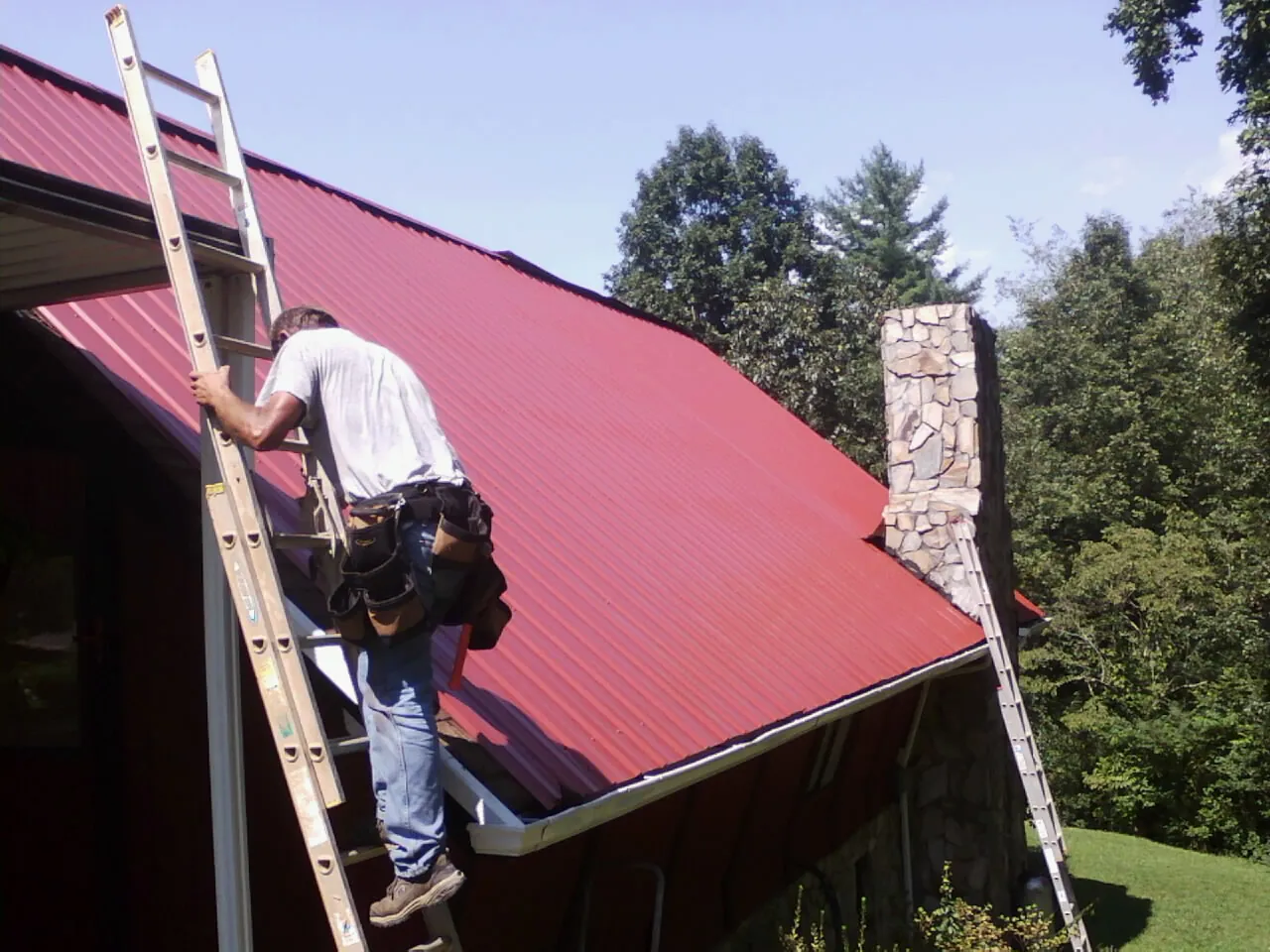 Roofer on a ladder beside a red metal roof and stone chimney — Soffit & Fascia Repair by Roofing Brothers in Pricedale
